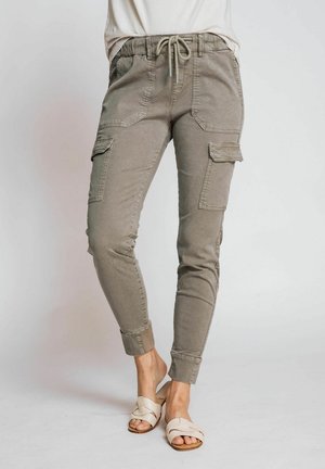 Khakifarbene Cargo-Hosen mit Kordelzug, schlanker Passform, mehreren Seitentaschen und weichem Stoff. Kombiniert mit beigen Slippern.