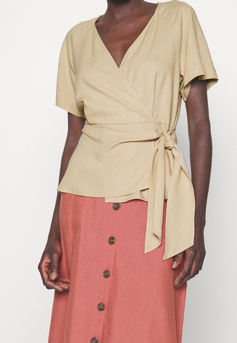 Personne portant un haut cache-cœur beige à manches courtes avec un lien sur le côté et une jupe rose taille haute boutonnée, debout devant un fond uni.