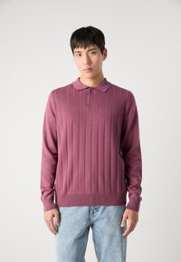 Pletena majica v utišani mauve barvi z navpično rebrasto teksturo, klasičnim ovratnikom in enim gumbom za zapiranje pri vratu. Nosi se z svetlo modrimi kavbojkami.