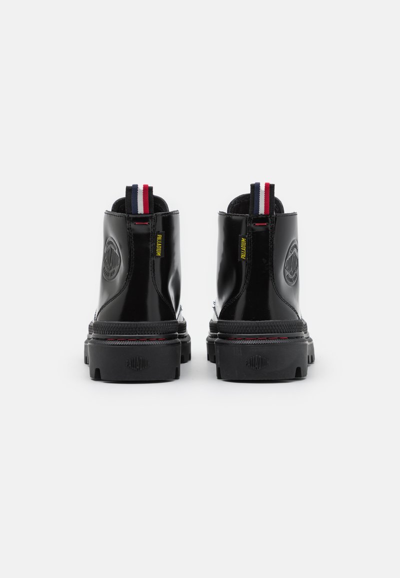 Bottines noires brillantes à bout rond, avec une semelle en caoutchouc texturée, un logo jaune et une tirette rayée rouge, blanche et bleue à l'arrière.