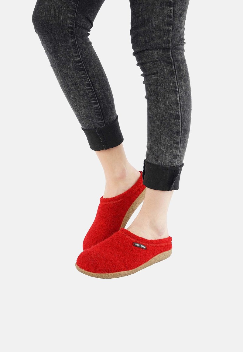 Personne portant un jean noir roulotté et des chaussons rouges à enfiler avec des semelles beiges, debout sur un fond blanc.