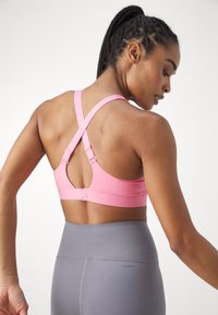 Roza športna modrček s križnimi naramnicami, nastavljivimi zapiranji, gladko teksturo in subtilnim sijajem. Uporabljen z visokim pasom sivih leggin.