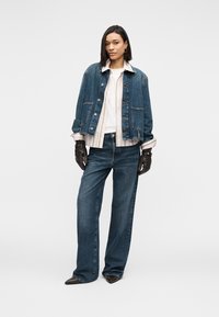 Veste en jean bleu foncé, associée à une chemise claire et un jean oversize. Le mannequin porte des gants noirs et des chaussures à bout pointu.