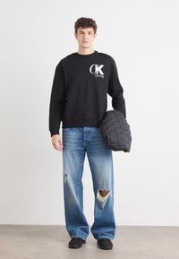 Hanorac negru din bumbac cu logo alb, asortat cu blugi lejera destramați de un albastru deschis, cu picior larg. Modelul ține o geantă neagră cu umăr, matlasată.