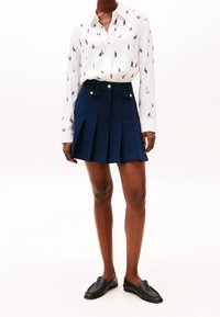 Chemise blanche à motifs de petits voiliers, assortie d'une jupe plissée bleu marine avec des boutons dorés et des poches. Des mocassins noirs viennent parfaire le look.