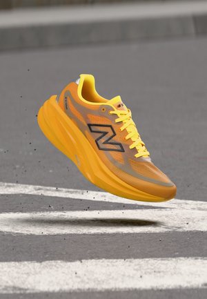 Une chaussure de course New Balance jaune orangé flotte au-dessus d’une piste en béton marquée de lignes blanches.