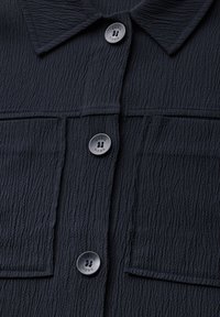 Camicia nera in tessuto testurizzato con colletto, due tasche frontali e bottoni rotondi con il logo "CECIL" impresso. Tessuto morbido e stropicciato.
