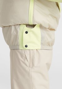 Beige pufferjack met een lichtgroene accent en zwarte drukknoopjes. Heeft een verstelbare tailleband en een zijzak met rits voor functionaliteit.