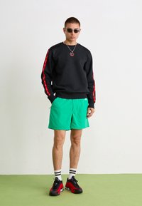Sort sweatshirt med røde sidelabber, grønne shorts, sorte og røde sneakers og solbriller. Enkle designs, casual pasform og livlige farver.