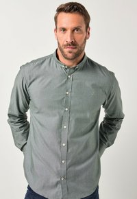 Camicia verde con colletto, caratterizzata da una leggera texture e un piccolo logo sul petto. I bottoni sono color crema. Maniche lunghe.