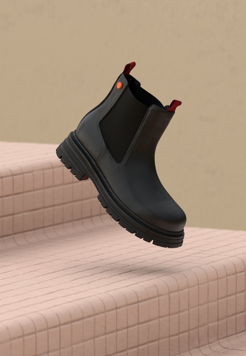 Botín Chelsea negro con paneles elásticos, lengüetas rojas y suela de goma texturizada. Acabado de cuero liso con detalles de costura mínimos.