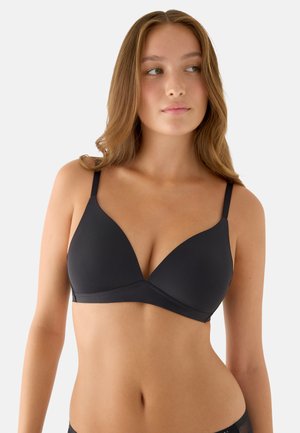 Mujer joven con cabello largo y castaño, vestida con un sujetador negro sencillo, mirando ligeramente hacia su derecha contra un fondo claro y liso.