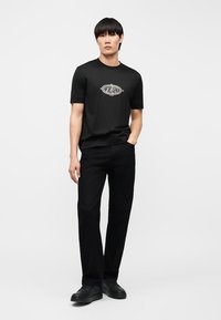 Άνδρας που στέκεται φορώντας μαύρο T-shirt με σχέδιο, μαύρο παντελόνι και μαύρα παπούτσια, μπροστά σε ένα απλό φωτεινό φόντο.