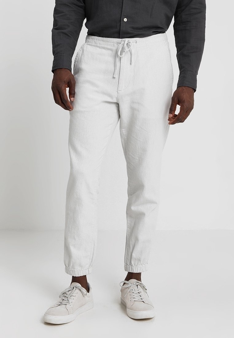 Man in witte joggers met trekkoord, beige sneakers en een zwarte button-up shirt, weergegeven van de taille naar beneden op een witte achtergrond.