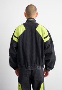 Person med rakad skalle klädd i svart och neon gult Adidas-jacka och matchande byxor, stående med ryggen mot en enfärgad bakgrund.