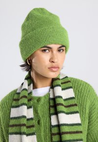 Berretto di maglia verde con una texture a coste, abbinato a un cardigan di maglia verde e a una sciarpa a righe con motivi verde e crema.