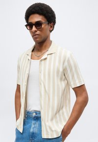 Άνδρας με κοντά, σγουρά μαλλιά που φοράει σκούρα γυαλιά ηλίου, μπλουζάκι με μπεζ και λευκές ρίγες με κοντά μανίκια πάνω από λευκό φανελάκι, μπλε τζιν και ασημένια αλυσίδα.