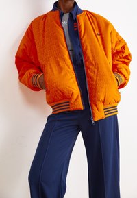 Orange quilted bomber jacket with a floral pattern, navy ribbed cuffs and hem. Worn over a navy zip-up and gray top.