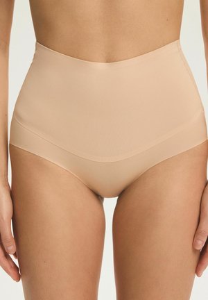 Bezszwowe cieliste figi modelujące z wysokim stanem noszone na dolnej części tułowia kobiety, z rękami swobodnie opuszczonymi wzdłuż boków.