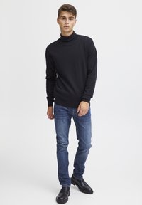 Svart genser med høy hals, lett strikket, kombinert med blå jeans. Modellen har på seg svarte snørestøvler. Enkel, tettsittende design, uten synlige mønstre.