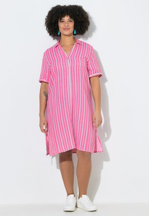 Femme debout portant une robe chemise à rayures roses et bleu clair avec manches courtes, des baskets blanches, des boucles d'oreilles turquoise et un tatouage sur le bras.