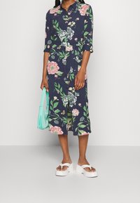 Abito a camicia floreale blu navy con fiori multicolori. Presenta maniche a tre quarti, cintura con laccio in vita e lunghezza midi. Indossato con sandali bianchi.