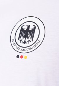 Λευκό T-shirt με στρογγυλό μαύρο λογότυπο που απεικονίζει έναν αετό και το κείμενο "Deutscher Handballbund", μαζί με μικρές μαύρες, κόκκινες και κίτρινες κουκκίδες.