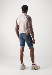 Helles pinkes und braunes Langarmoberteil für Herren mit gerippten Bündchen, kombiniert mit blauen Jeansshorts und beigen Turnschuhen mit gestreiften Socken.