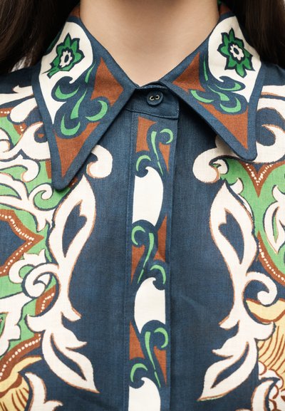 Κοντινό πλάνο για τον γιακά πουκάμισου με κουμπιά, διακοσμημένο με πολύπλοκα πράσινα, καφέ και λευκά floral και στροβιλιστά σχέδια σε σκούρο μπλε ύφασμα.