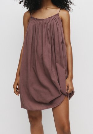 Femme portant une robe mauve ample et sans manches avec un col plissé, debout devant un fond blanc uni.