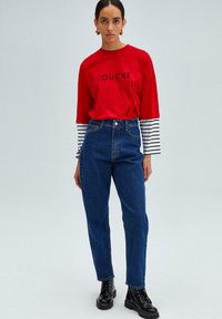 Camisa roja de manga larga con letras negras, puños a rayas; jeans oscuros de cintura alta; botas negras con cordones. Colores sólidos, ajuste casual.