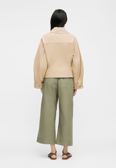 Γυναίκα με μακριά σκούρα μαλλιά που φοράει μπεζ μπουφάν, ελιά φαρδιά κατεστραμμένα παντελόνια και καφέ σανδάλια με λουράκια, στέκεται γυρισμένη πλάτη σε λευκό φόντο.