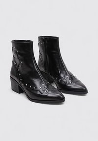 Botas de cuero negras hasta el tobillo con punteras afiladas, tachuelas metálicas y un tacón cuadrado. Cuentan con un cierre de cremallera y un diseño de costura decorativa.