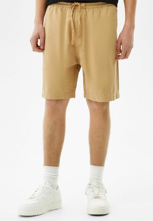 Homme portant un short beige avec cordon, des chaussettes blanches et des baskets blanches, debout devant un fond blanc.