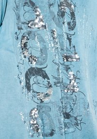 Hellblaues T-Shirt mit metallisch silbernen Pailletten in einem grafischen Design angeordnet, ergänzt durch kontrastierenden dunkelbedruckten Text und abstrakte Muster.
