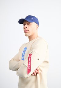 Μπεζ φούτερ με μπλε λογότυπο, τσέπη με φερμουάρ στο πλάι και κόκκινη ετικέτα "Αφαιρέστε Πριν την Πτήση". Το ναυτικό καπέλο ολοκληρώνει την εμφάνιση.