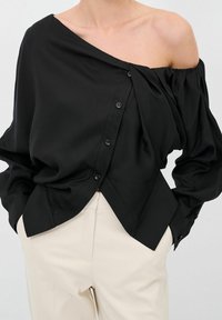 Blusa nera a spalle scoperte con vestibilità ampia, caratterizzata da una chiusura a bottoni su un lato e delicate drappeggiature. Tessuto dalla consistenza liscia.