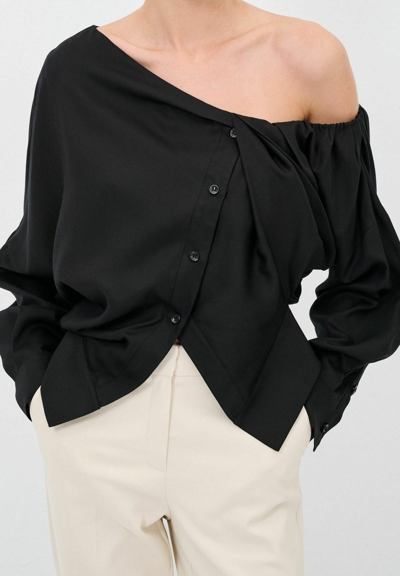 Blusa nera a spalle scoperte con vestibilità ampia, caratterizzata da una chiusura a bottoni su un lato e delicate drappeggiature. Tessuto dalla consistenza liscia.