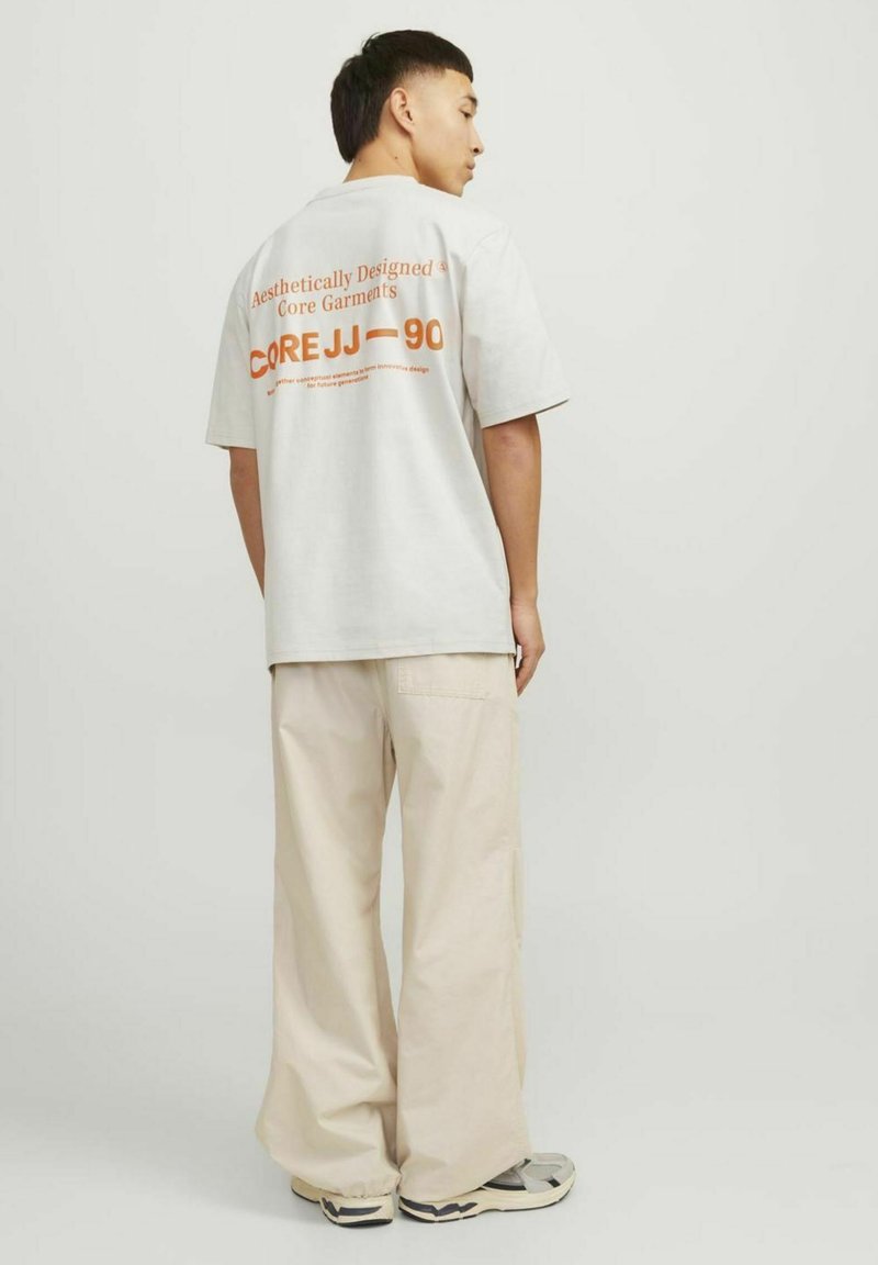 Light gray oversized t-shirt with orange text on the back, paired with beige wide-leg pants and gray sneakers. Simple, casual design.