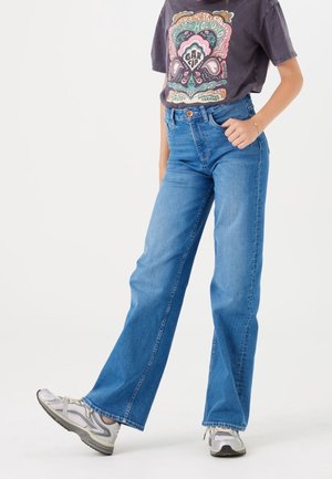 Person iført blå højttaljede vidde bukser, grafisk t-shirt og hvide sneakers, stående med den ene fod let hævet.
