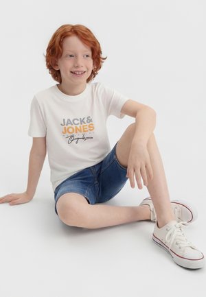 Rothaariger Junge sitzt entspannt auf dem Boden, trägt ein weißes Jack & Jones T-Shirt, Jeansshorts und weiße Sneakers mit Schnürsenkeln.