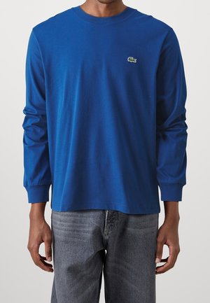 Homme portant une chemise bleu à manches longues avec un petit logo crocodile vert sur la poitrine et un jean gris.