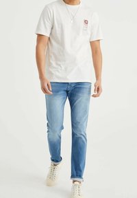 Man som bär en vit T-shirt med kort ärm och en liten röd grafik, ljusblå jeans, vita sneakers och ett silverhalsband.