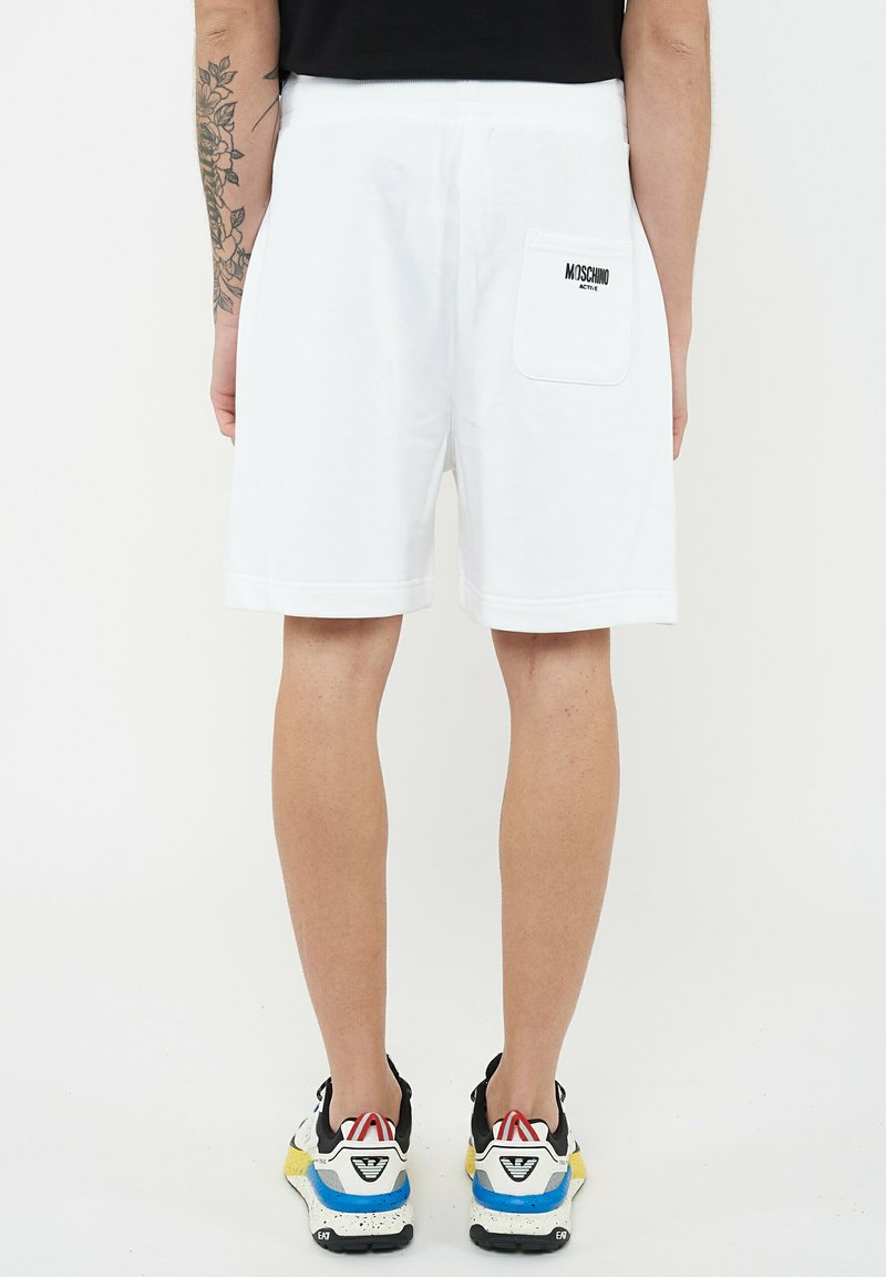 Homme portant un short blanc Moschino Active avec une poche arrière et des baskets noires, bleues, jaunes et blanches, debout devant un fond uni.
