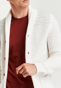 White knitted cardigan with a textured pattern and shawl collar, featuring wooden buttons, worn over a maroon t-shirt.