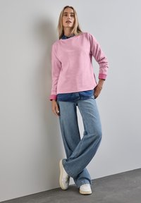 Maglione rosa a maniche lunghe sopra una camicia di denim, abbinato a jeans blu a gamba larga e scarpe da ginnastica bianche, su uno sfondo grigio.