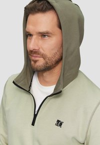 Lichtgroene ritshoodie met een contrasterende olijfgroene capuchon. Voorzien van een zwarte ritssluiting en logo op de borst. Glad textuur, casual ontwerp.