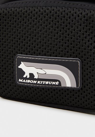 Sac en maille noire avec un patch rectangulaire présentant un logo de renard et des rayures en tons de gris, étiqueté "MAISON KITSUNÉ." Des surfaces texturées sont visibles.