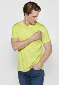 Tricou galben strălucitor cu mânecă scurtă, guler rotund și logo subtil imprimat pe piept. Materialul pare moale și ușor.
