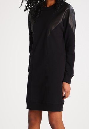 Nainen, joka pukeutuneena mustaan polven mittaiseen neulemekkoon, jossa on nahkapaneelit hartioissa ja pitkät hihat, vaaleaa tasaista taustaa vasten.